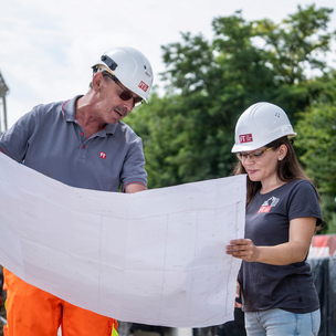 Zwei Mitarbeiter der UNTERNEHMENSGRUPPE KASSECKER Ein älterer Mann und eine junge Frau halten einen Baustellenplan, beide tragen einen Bauhelm-