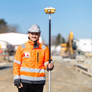 Bauzeichner Auszubildender von KASSECKER bei der Vermessung auf einer Baustelle Ein Bauarbeiter in orangefarbener Sicherheitskleidung und Helm steht auf einer Baustelle und hält eine Vermessungsausrüstung. Im Hintergrund sind Baumaterialien und Baustellenfahrzeuge zu sehen.