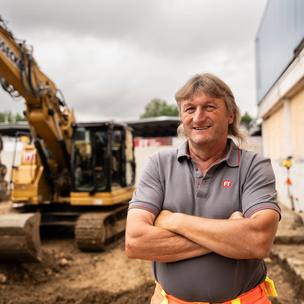 Baustellenpersonal Maschinist von Tausendpfund Freundlicher älterer Mann mit verschränkten Armen steht vor einem großen Bagger auf einer Baustelle.