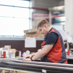 KASSECKER Auszubildender zum Metallbauer bei der Arbeit in der Werkstatt Ein junger Mann in Arbeitskleidung und Schutzweste arbeitet in einer Werkstatt an einem Metallrahmen. Im Hintergrund sind weitere Werkzeuge und Materialien zu sehen.