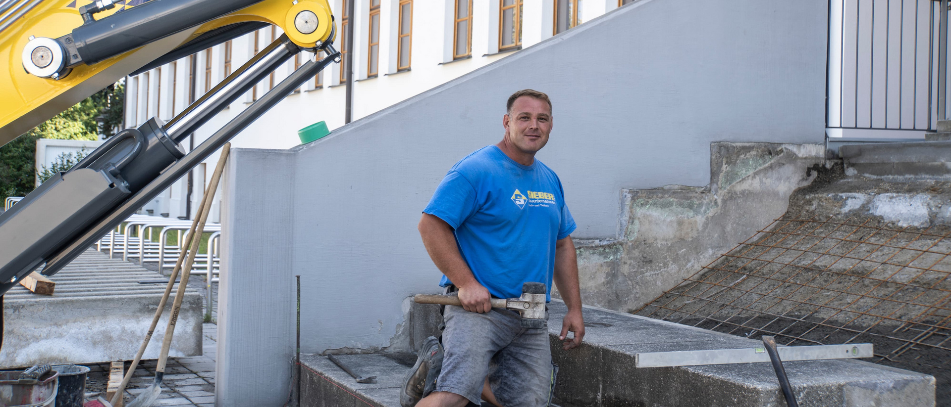 Ein Mann kniet auf einer Treppe und hält einen Gummihammer. Im Hintergrund ein Bagger.