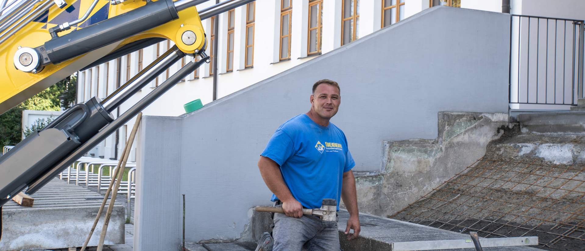 Ein Mann kniet auf einer Treppe und hält einen Gummihammer. Im Hintergrund ein Bagger.