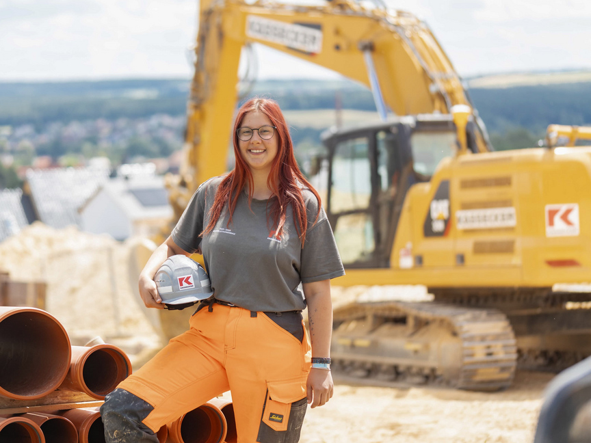 Auszubildende der Unternehmensgruppe Kassecker zur Rohrleitungsbauerin Eine junge Frau in Arbeitskleidung und Helm hält einen Bauhelm und steht neben großen Rohren auf einer Baustelle. Im Hintergrund sind Baugeräte und Erdhaufen zu sehen.