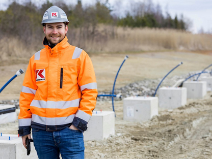 KASSECKER Mitarbeiter Manuel Ein lächelnder KASSECKER Mitarbeiter in orangefarbener Sicherheitskleidung und Helm steht auf einer Baustelle. Im Hintergrund sind Betonblöcke und Rohre zu sehen.