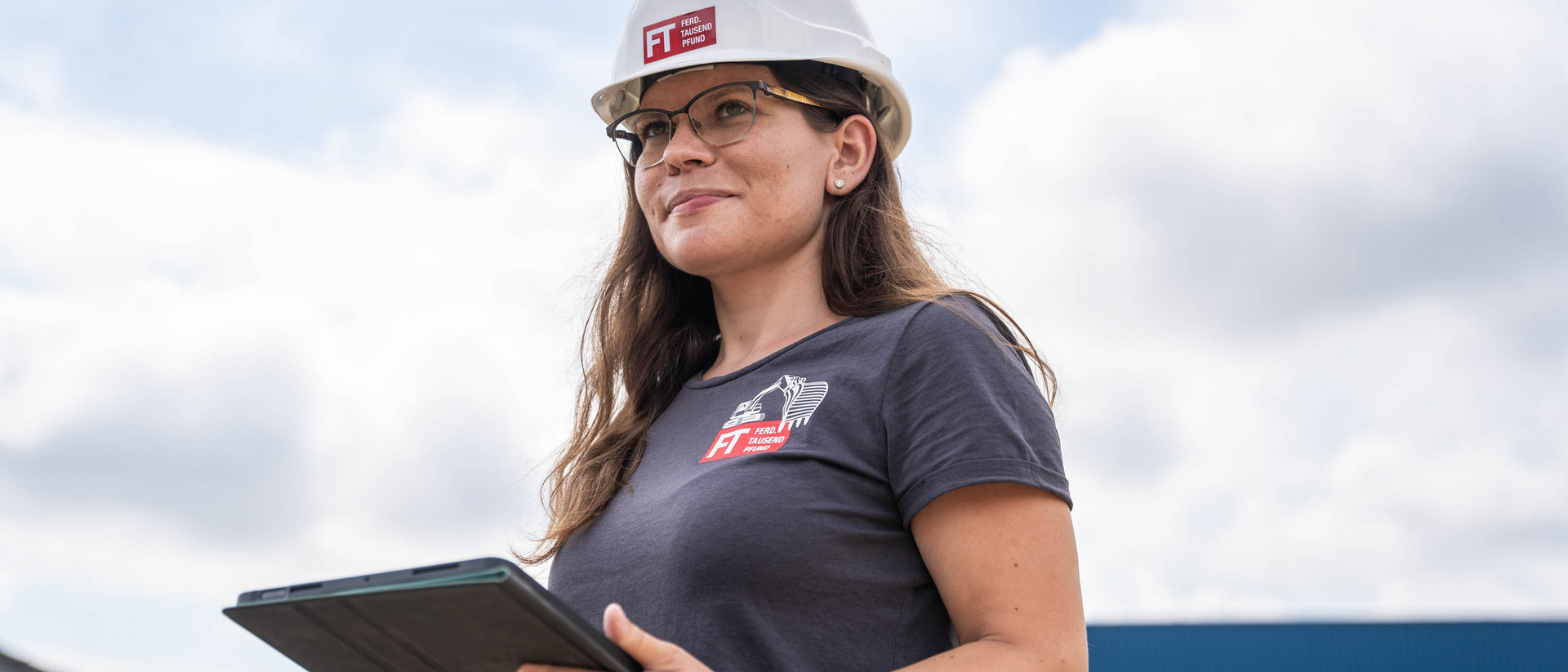 Mitarbeiterin mit Bauhelm steht mit Tablet auf Baustelle und lächelt freundlich in die Ferne.