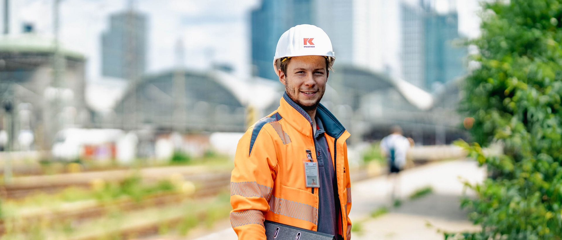 Bauarbeiter in auffälliger Kleidung hält ein Klemmbrett neben Eisenbahnschienen, mit Industrieanlagen und Wolkenkratzern im Hintergrund.