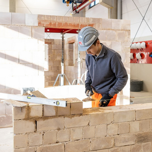 Auszubildender zum Maurer der Unternehmensgruppe Kassecker beim Mauern einer Wand Ein Auszubildender in Arbeitskleidung und Helm mauert eine Wand in einer Werkstatt. Werkzeuge und Baumaterialien sind um ihn herum zu sehen.
