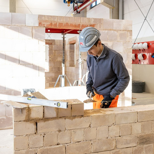 Auszubildender zum Maurer der Unternehmensgruppe Kassecker beim Mauern einer Wand Ein Auszubildender in Arbeitskleidung und Helm mauert eine Wand in einer Werkstatt. Werkzeuge und Baumaterialien sind um ihn herum zu sehen.