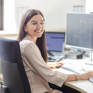 Industriekauffrau Auszubildende bei der Büroarbeit Eine junge Frau mit Brille sitzt an einem Schreibtisch in einem Büro und arbeitet an einem Computer. Sie lächelt in die Kamera.