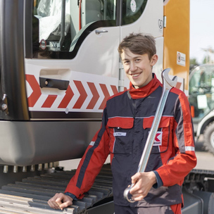 Auszubildender Land- und Baumaschinenmechatroniker bei der Arbeit Ein junger Mann in Arbeitskleidung steht mit einem großen Schraubenschlüssel vor einer Baumaschine und lächelt in die Kamera.