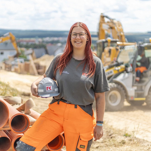 Auszubildende Rohrleitungsbauerin bei der Arbeit auf der Baustelle Eine junge Frau in Arbeitskleidung und Helm hält einen Bauhelm und steht neben großen Rohren auf einer Baustelle. Im Hintergrund sind Baugeräte und Erdhaufen zu sehen.