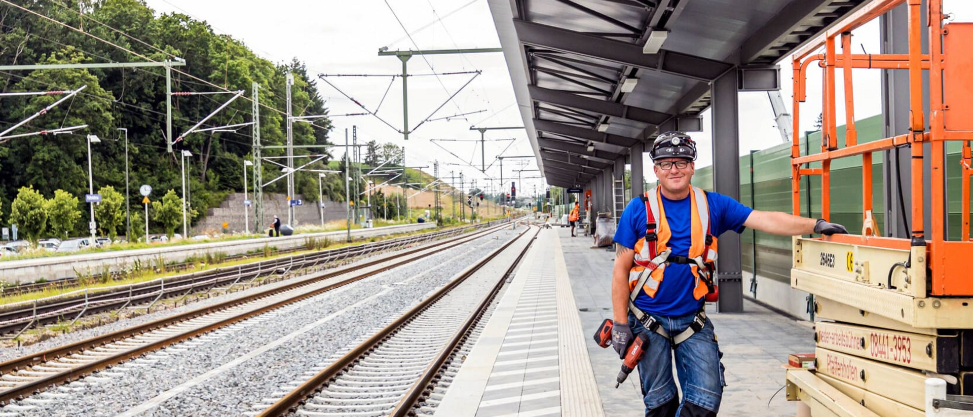 Ein Bauarbeiter mit Sicherheitsausrüstung steht auf einem Bahnsteig neben einer orangefarbenen Arbeitsbühne. Im Hintergrund sind Gleise und eine Überdachung zu sehen.