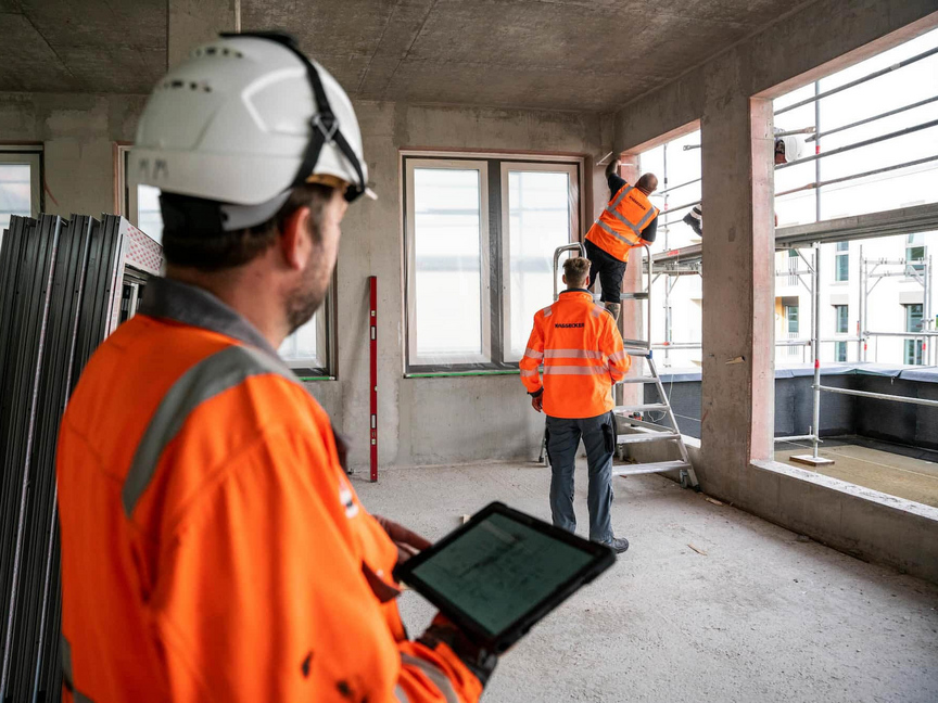 Stahl und Metallbau bei KASSECKER Bauarbeiter in orangefarbener Arbeitskleidung von KASSECKER arbeiten im Innenausbau eines Gebäudes. Einer der Arbeiter steht auf einem Gerüst, um Messungen vorzunehmen und ein anderer nutzt ein Tablet.